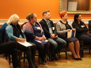 Grupa osób siedzi na krzesłach w jednym rzędzie przy ścianie. Wszyscy patrzą w jednym kierunku.