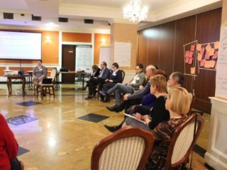 Grupa osób siedzi na krzesłach w pół okręgu i patrzy w kierunku osoby prowadzącej i prezentacji multimedialnej.