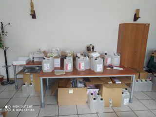 Stół zastawiony kartonami oraz pięciolitrowymi plastikowymi kanistrami z cieczą. Pod stołem i obok też stoją kanistry i kartony.