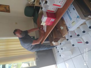 Mężczyzna w maseczce podpisuje dokument który leży na stole na którym i pod którym stoją plastikowe kanistry z cieczą.