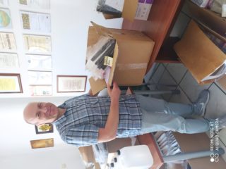 Mężczyzna stoi przy stołach i trzyma pełny karton który stoi na stole. Z tyłu stoi stół z kartonami i plastikowymi kanistrami.