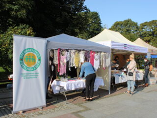 Trzy stoiska z upominkami stoją w jednym rzędzie na tle zielonych drzew. Przy stoiskach stoi kilka osób.