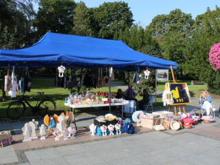 Niebieskie stoisku z drewnianymi aniołkami, koszykami, torbami. Obok stoi tablica z żółto-czarnym liściem klonu. Obok siedzi kilka osób.