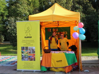 Żółte niewielkie stoisko KLANZA za którym stoją dwie osoby a obok stoi baner. Na stoliku znajdują się ulotki i wiklinowy kosz.