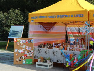 Stoisko MOPS Łapy na którym znajdują się zdobione butelki, słoiki, balony, małe rośliny doniczkowe, laurki. Obok stoi obraz.