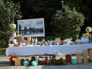 Stół z białym obrusem zastawiony rękodziełem takim jak obrazki, koszyki, kwiaty, misy. W tle stoi czarno-biały obraz a za nim widać zielony park.