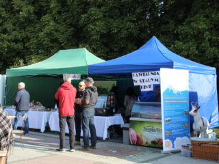 Dwa stoiska jedno obok drugiego na tle zielonych drzewa. Jedno ze stoisk sprzedaje kawę. Wokół stoi kilka osób.