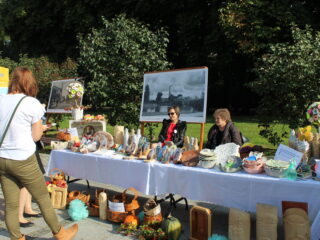 Dwie kobiety siedzą za stołem z białym obrusem na którym znajdują się rękodzieła typu obrazki, koszyki, kwiaty, misy. Z tyłu stoją dwa duże obrazy a dalej widać zielony park.