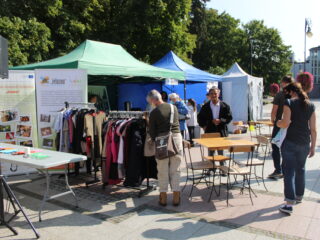 Trzy stoiska przed którymi stoją stoliki z krzesłami. Obok stoisk znajdują się stojaki z ubraniami które oglądają przechodni.
