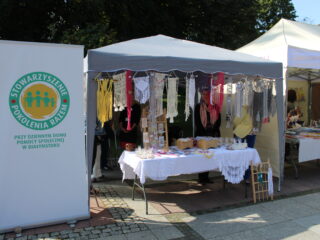 Stoisko z ręcznie dzierganymi ozdobami oraz z wiklinowymi koszykami. Obok stoi baner Stowarzyszenia Pokolenia Razem.