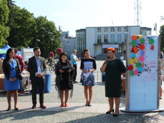 Grupa osób stoi obok kobiety która przemawia przez mikrofon na środku placu przed Teatrem Dramatycznym.