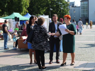 Trzy osoby wręczają kobiecie podarunek. W dalszym planie widać stoiska i ludzi chodzących obok.