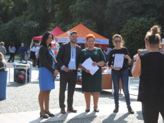 Cztery uśmiechnięte osoby na tle stoisk pozują do zdjęcia robionego przez kobietę przy pomocy telefonu.