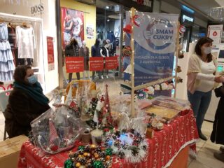 Stoisko z ozdobami bożonarodzeniowymi typu martwa natura, małe choinki, bombki. Przy stoisku siedzi kobieta w maseczce.
