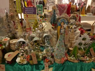 Stół z ozdobami bożonarodzeniowymi typu małe sztuczne choinki, aniołki oraz ozdoby z martwej natury.