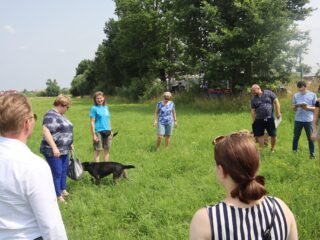 Grupa osób stoi w kółku na zielonej polanie. Miedzy nimi chodzi pies.