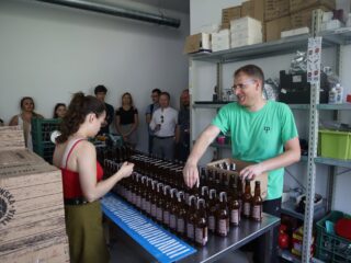 Grupa osób stoi przy ścianie i patrzy w stronę dwóch osób które stoją przy stole z rzędami ciemnych, szklanych butelek.