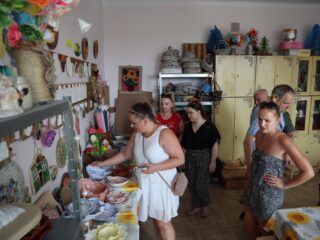 Grupa osób która ogląda wystawę z rękodziełem typu misy, talerze, sztuczne kwiaty, koszyki. W pomieszczeniu stoi duża szafa.