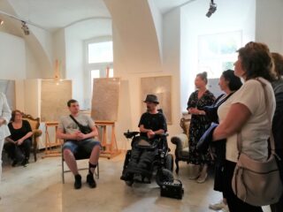 Grupa osób oraz mężczyzna na elektrycznym wózku inwalidzkim. Z tyłu na sztalugach stoją obrazy.