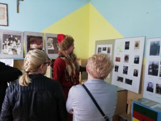 Mężczyzna w czerwonym stroju i futrze z lisa oprowadza ludzi po sali z historycznymi zdjęciami.