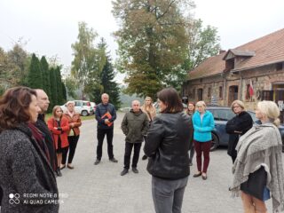 Grupa osób stoi na parkingu wokół osoby prowadzącej. Obok stoi stary, murowany budynek. W tle widać zielone drzewa.