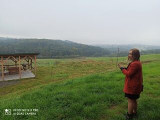 Kobieta w czerwonym płaszczu stoi na przeciwko pustej zielonej przestrzeni. Kawałek dalej stoi altanka z ławką. W oddali widać gęste lasy.