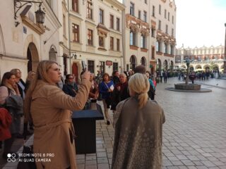 Grupa osób stoi na uliczce miasta pośród zabytkowych budynków. Na środku uliczki stoi fontanna.