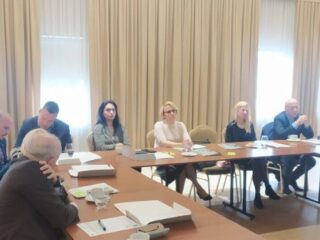 Sala z grupą osób która siedzi przy stołach ustawionych w kształt litery C. Na stołach leżą dokumenty.