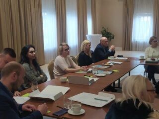 Grupa osób siedzi przy stołach ustawionych w kształt litery C. Na stołach leżą dokumenty. Większość patrzy w jednym kierunku.