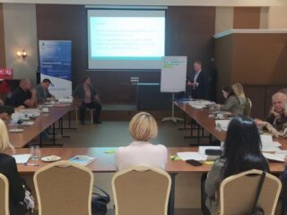 Sala z grupą osób która siedzi przy stołach ustawionych w kształt litery U na przeciwko prezentacji. Na stołach leżą dokumenty.