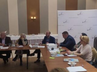 Grupa osób siedzi przy łączonych stołach na których leżą dokumenty. Wszyscy patrzą w jednym kierunku. W tle widać stół z poczęstunkiem.