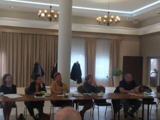 Grupa osób siedzi przy łączonych stołach na których leżą dokumenty. Z tyłu widać pustą przestrzeń sali oraz drzwi.