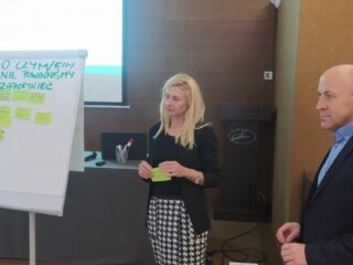 Kobieta i mężczyzna stoją przy tablicy z notatkami i przyklejonymi karteczkami. Kobieta trzyma zieloną karteczkę. Z tyłu widać ekran projekcyjny z wyświetloną prezentacją.