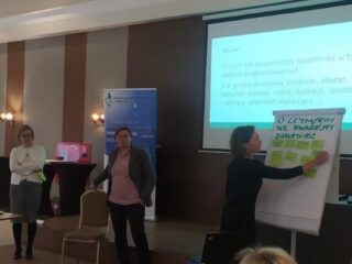 Kobieta notuje na tablicy. Obok przy stole z resztą uczestników stoją jeszcze dwie kobiety. W tle jest wyświetlona prezentacja.