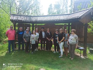 Stowarzyszenie Muzeum Małej Ojczyzny w Studziwodach. Grupowe zdjęcie na tle drewnianych tablic pod daszkiem.
