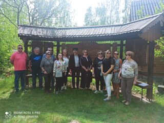 Stowarzyszenie Muzeum Małej Ojczyzny w Studziwodach. Grupowe zdjęcie na tle drewnianych tablic pod daszkiem.