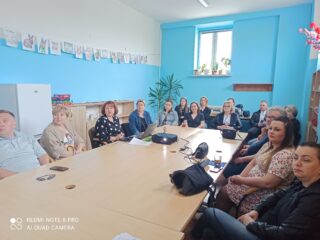 Centrum Integracji Społecznej w Hajnówce. Grupa osób siedzi przy dużym stole i ogląda wyświetloną prezentację.