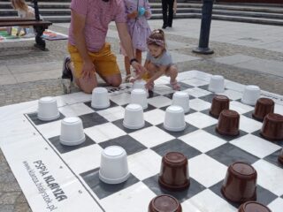 11 Podlaskie Targi Ekonomii Społecznej w Białymstoku na Rynku Kościuszki. Mężczyzna oraz dwie dziewczynki grają w warcaby na placu na dużej planszy.