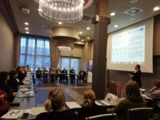 Spotkanie Regionalnego Komitetu Ekonomii Społecznej w hotelu 3 Trio. Uczestnicy siedzą przy łączonych stołach ustawionych w kształt litery C. Obok ekranu projekcyjnego z wyświetloną prezentacją stoi kobieta która przemawia.