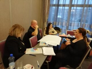 Spotkanie Regionalnego Komitetu Ekonomii Społecznej w hotelu 3 Trio. Cztery osoby siedzą przy podłużnych łączonych stołach i rozmawiają. Na stole leżą teczki oraz dokumenty.