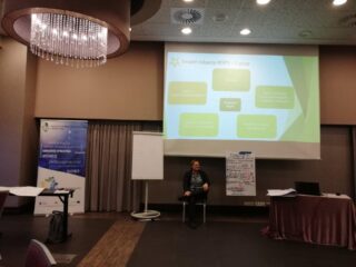 Spotkanie Regionalnego Komitetu Ekonomii Społecznej w hotelu 3 Trio. Kobieta siedzi pod ekranem projekcyjnym z wyświetloną prezentacją. Obok stoi stolik z laptopem, tablica oraz przyklejona do ekranu projekcyjnego duża kartka z notatkami.