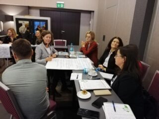 Spotkanie Regionalnego Komitetu Ekonomii Społecznej w hotelu 3 Trio. Grupa osób siedzi przy podłużnych łączonych stołach z czego jedna z osób pisze na dużej kartce. Na stole leżą teczki oraz dokumenty. Na drugim planie widać kolejną pracującą grupę.