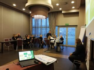 Spotkanie Regionalnego Komitetu Ekonomii Społecznej w hotelu 3 Trio. Grupa osób siedzi przy łączonych stołach w kształcie litery L. Jedna z siedzących kobiet trzyma dwie duże kartki. Obok ekranu projekcyjnego siedzi kobieta a obok niej stoi stolik z laptopem.