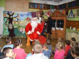 Dzieci siedzą na podłodze przed świętym Mikołajem. Za plecami Mikołaja stoją dekoracje, dwie kobiety oraz dziewczynka.