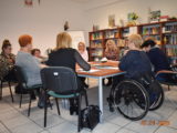 Grupa kobiet siedzi przy stole z dokumentami. Jedna z uczestniczek siedzi na wózku inwalidzkim.