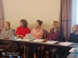 Warsztaty sieciujące nt. postępowania wobec osób z zachowaniami autodestrukcyjnymi. Cztery kobiety oraz jeden mężczyzna siedzą przy podłużnym stole i wszyscy patrzą w bok. Każdy ma przed sobą teczkę z dokumentami.
