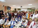 Trzecie spotkanie pt. Forum Inicjatyw Lokalnych w woj. lubelskim. Liczne grono uśmiechniętych osób w ludowych strojach pozuję do grupowego zdjęcia na dużej sali. Większość stoi w półokręgu a na środku kilkanaście osób siedzi na podłodze.